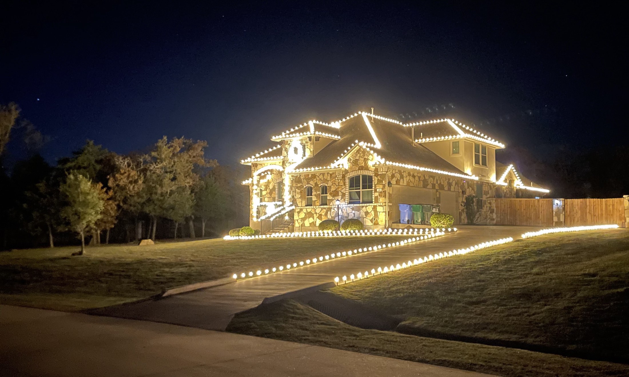 A two-story stone home at night outlined in warm white roofline lighting, with lit stakes along the driveway