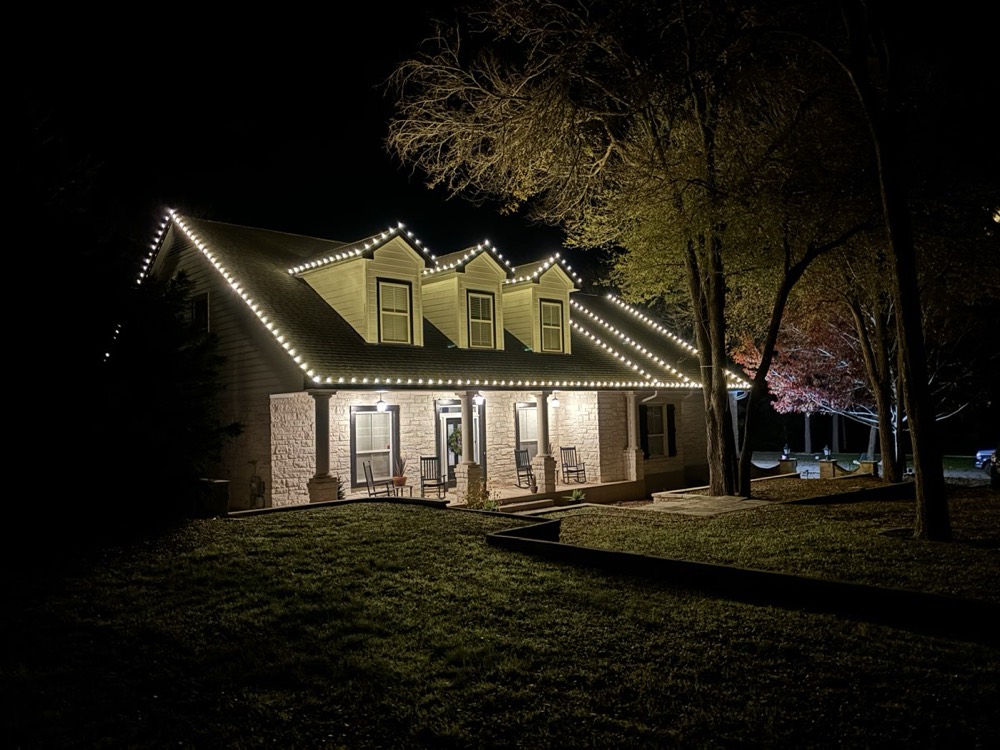 A Hill Country farmhouse at night with warm white lights tracing the roofline, three dormer peaks, and the front porch
