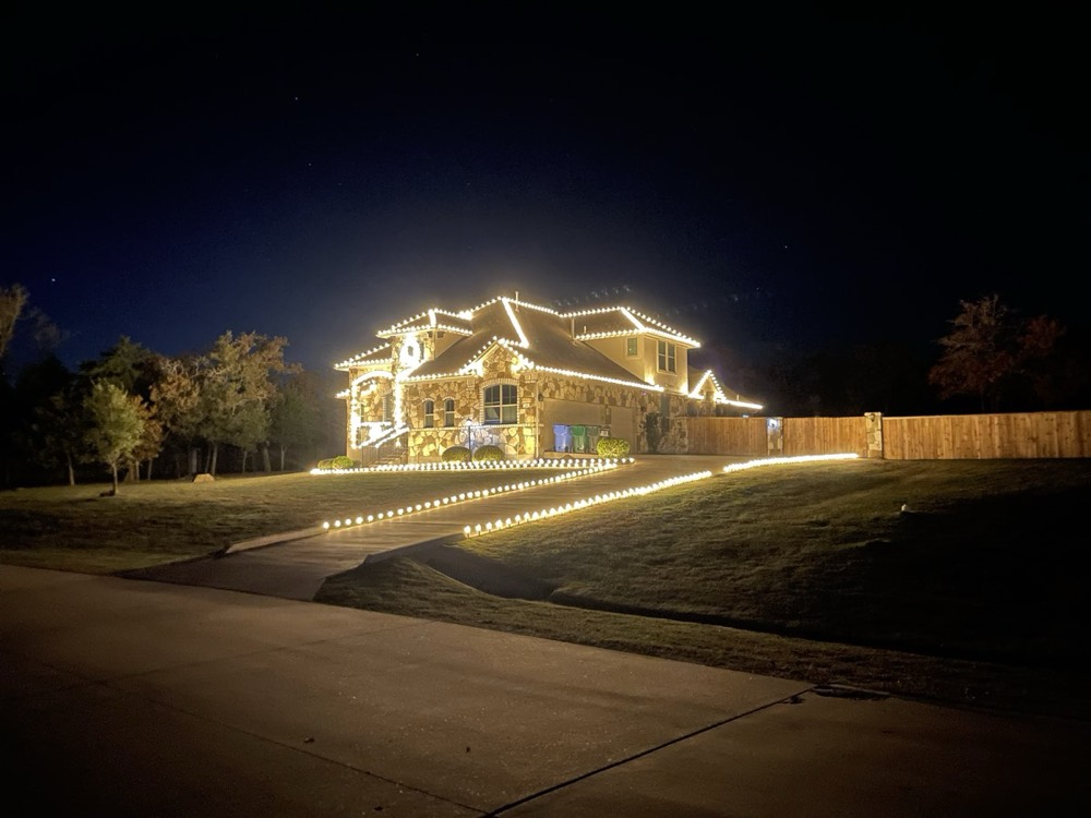 A two-story stone home outlined in warm white lighting, with lit stakes running along the driveway and walkway