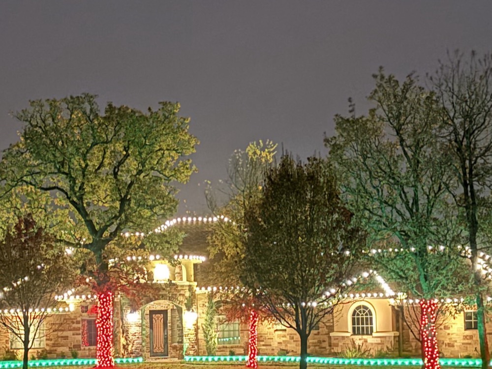 A stone ranch home lit in red and green, with white roofline lights, red-wrapped tree trunks, and a green-lit border along the beds
