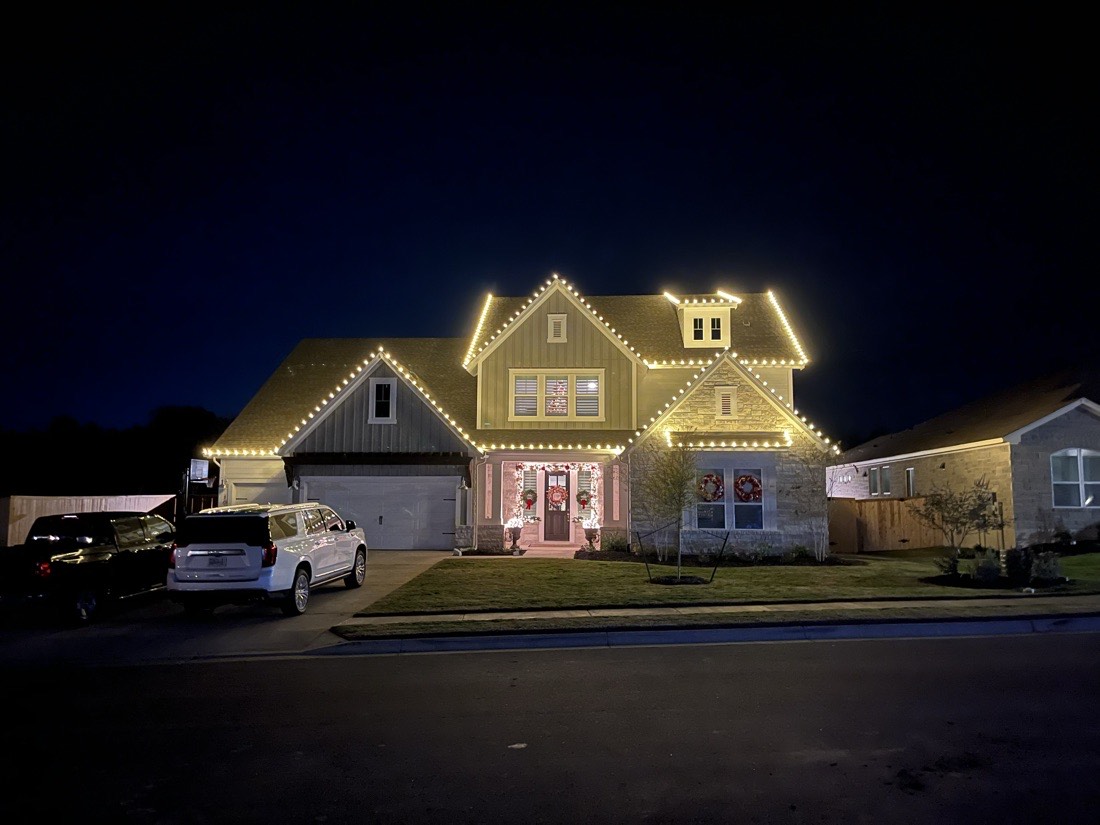 A two-storey home at night with warm white lights along every gable and dormer, wreaths on the upper windows, and lit porch columns